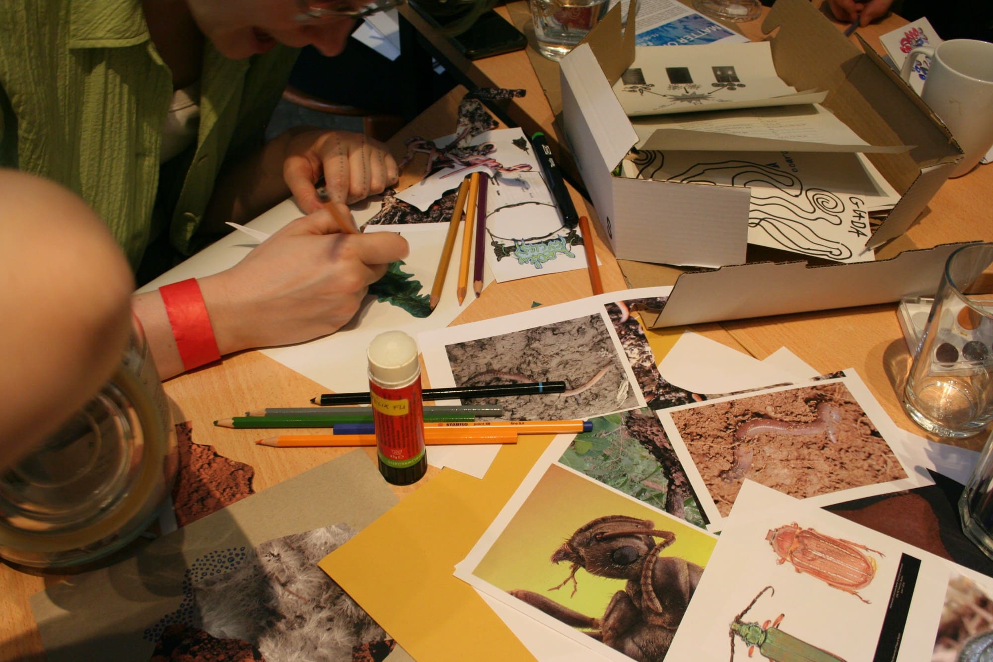 someone drawing at a table with lots of drawings, photographs paper, pencils a stick of glue