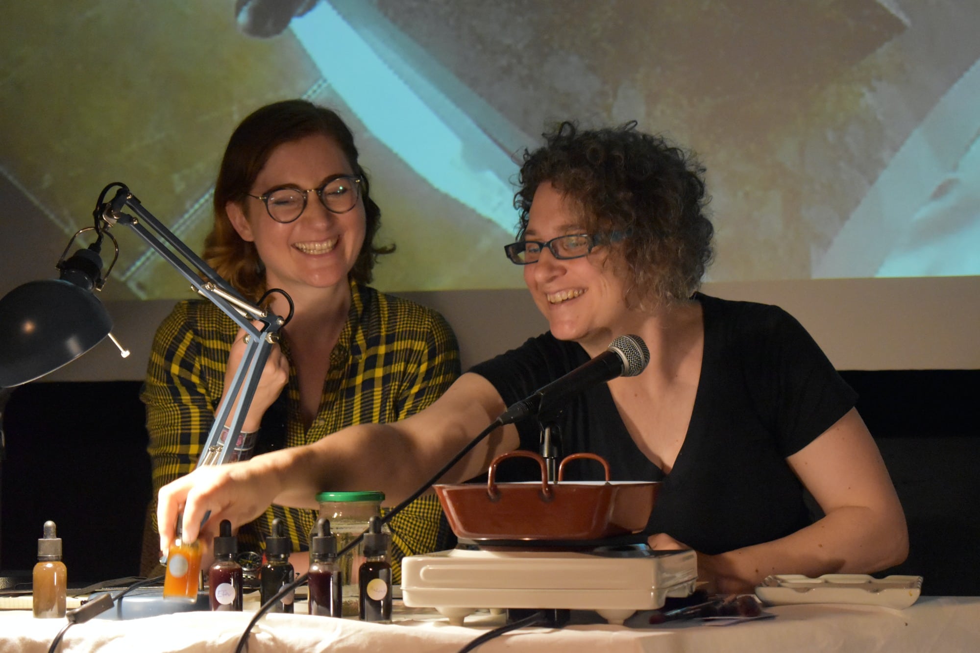 two people seated laughing, one gabs a bottle with yellow liquid