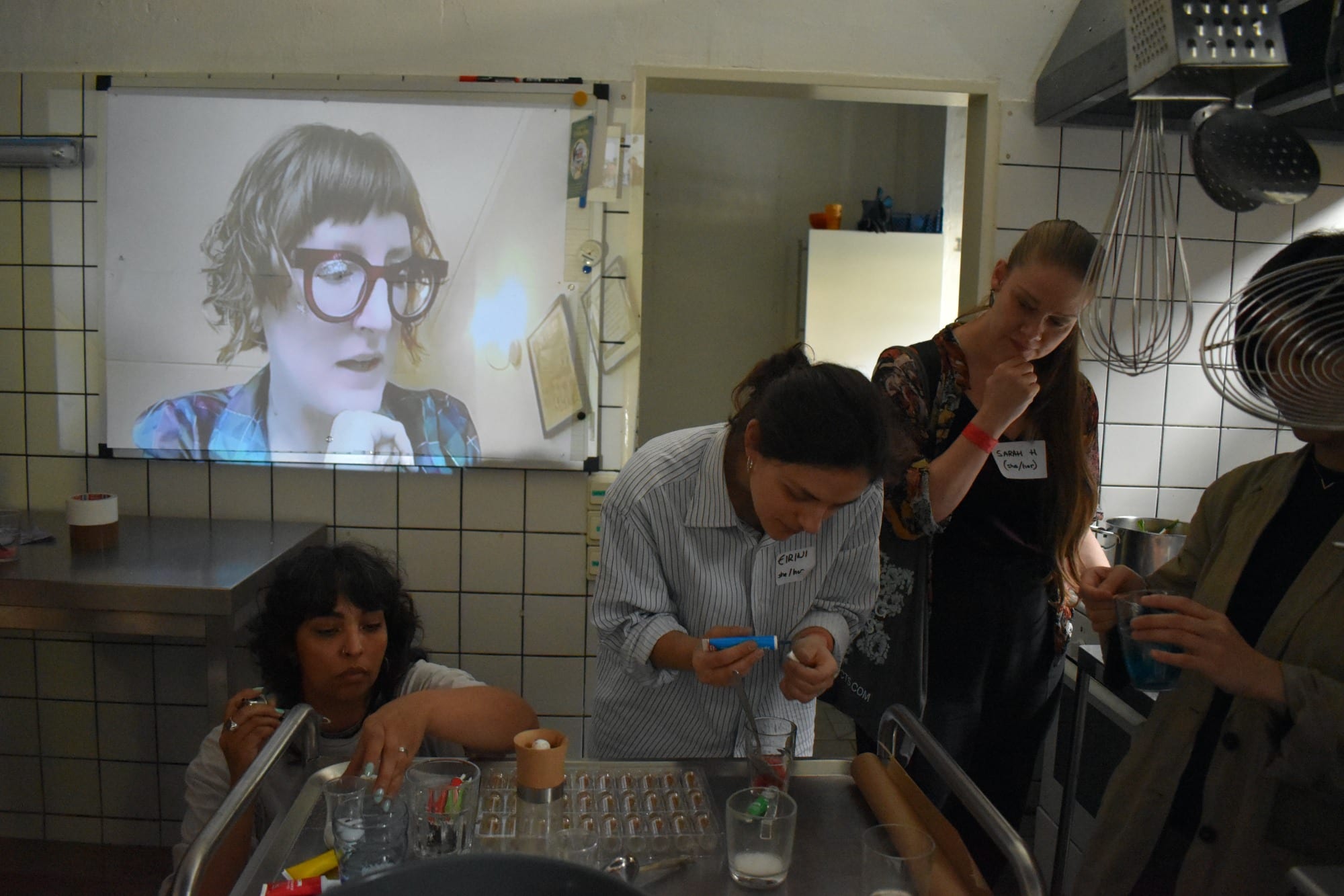 Three women working around a metal table a projection on the wall show the head of another woman.