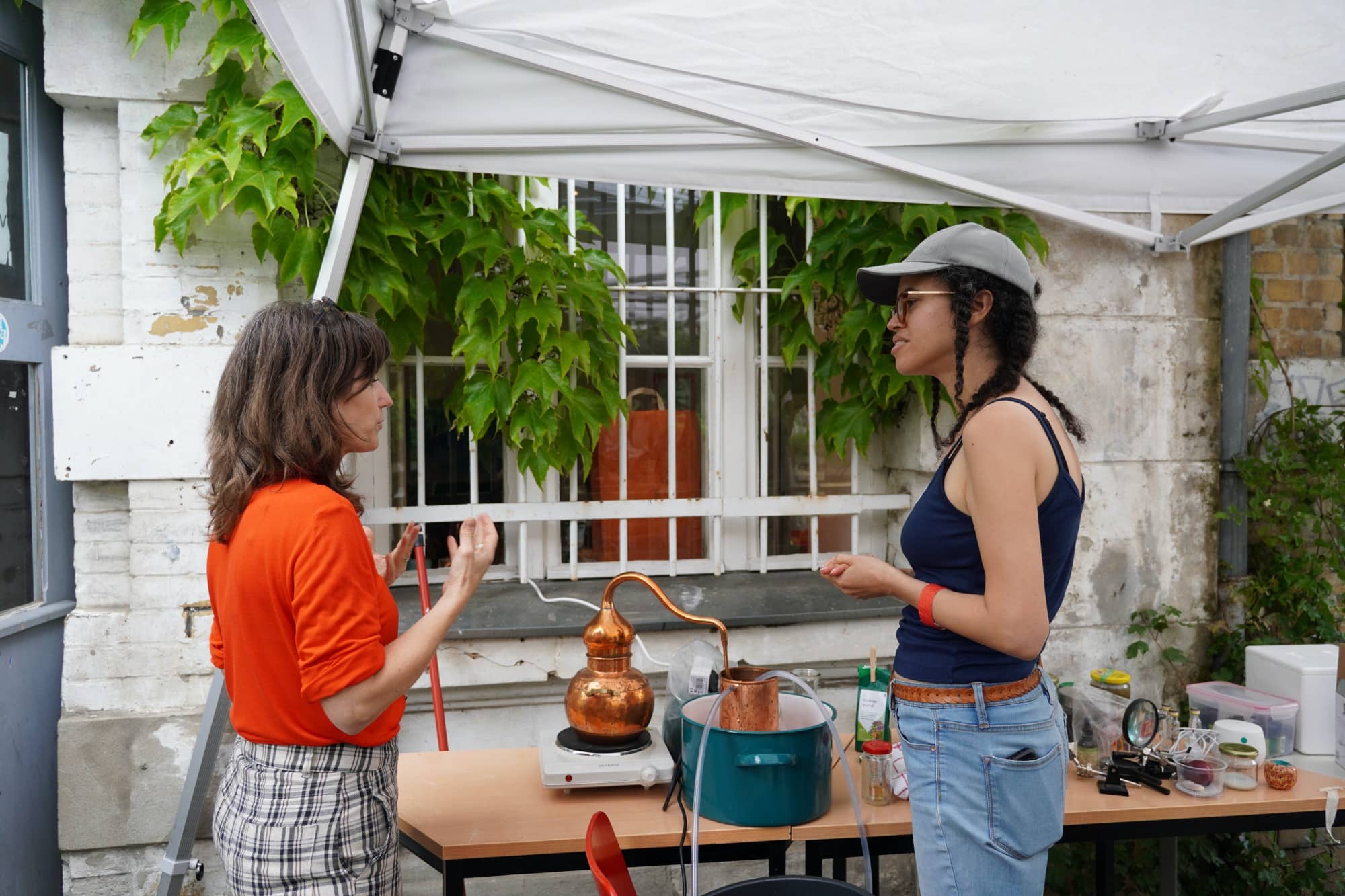 two women standing outside a building. a brass distiller sits on a table 