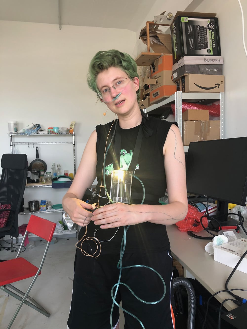 Photo of an artist with green hair and a tube between a container and her nose