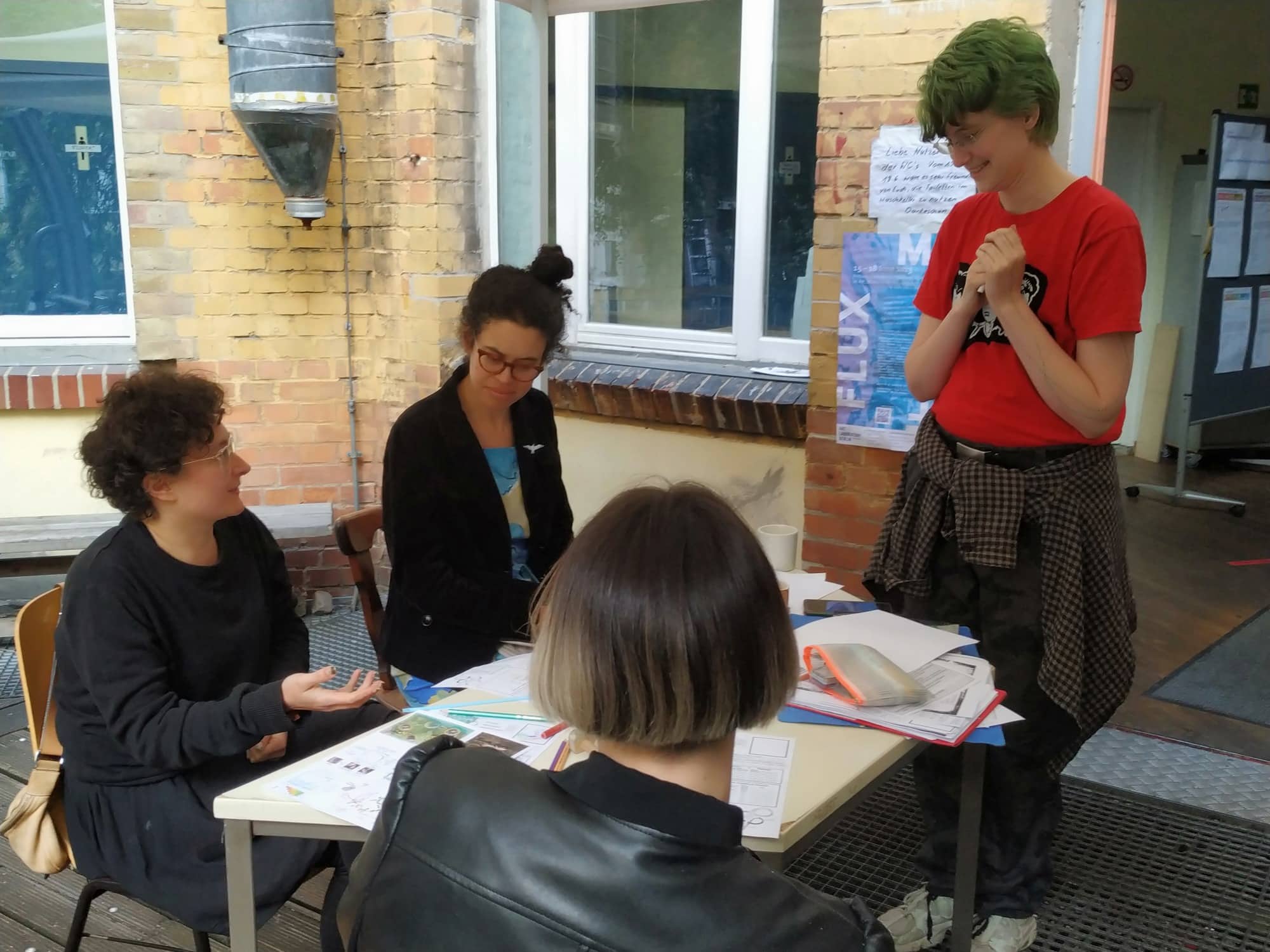 three women seated at a table outside a building, a fourth stands and smiles