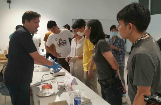 A man with blue glove stands on one side of a table with laboratory equipment talking to a group of people on the other side of the table
