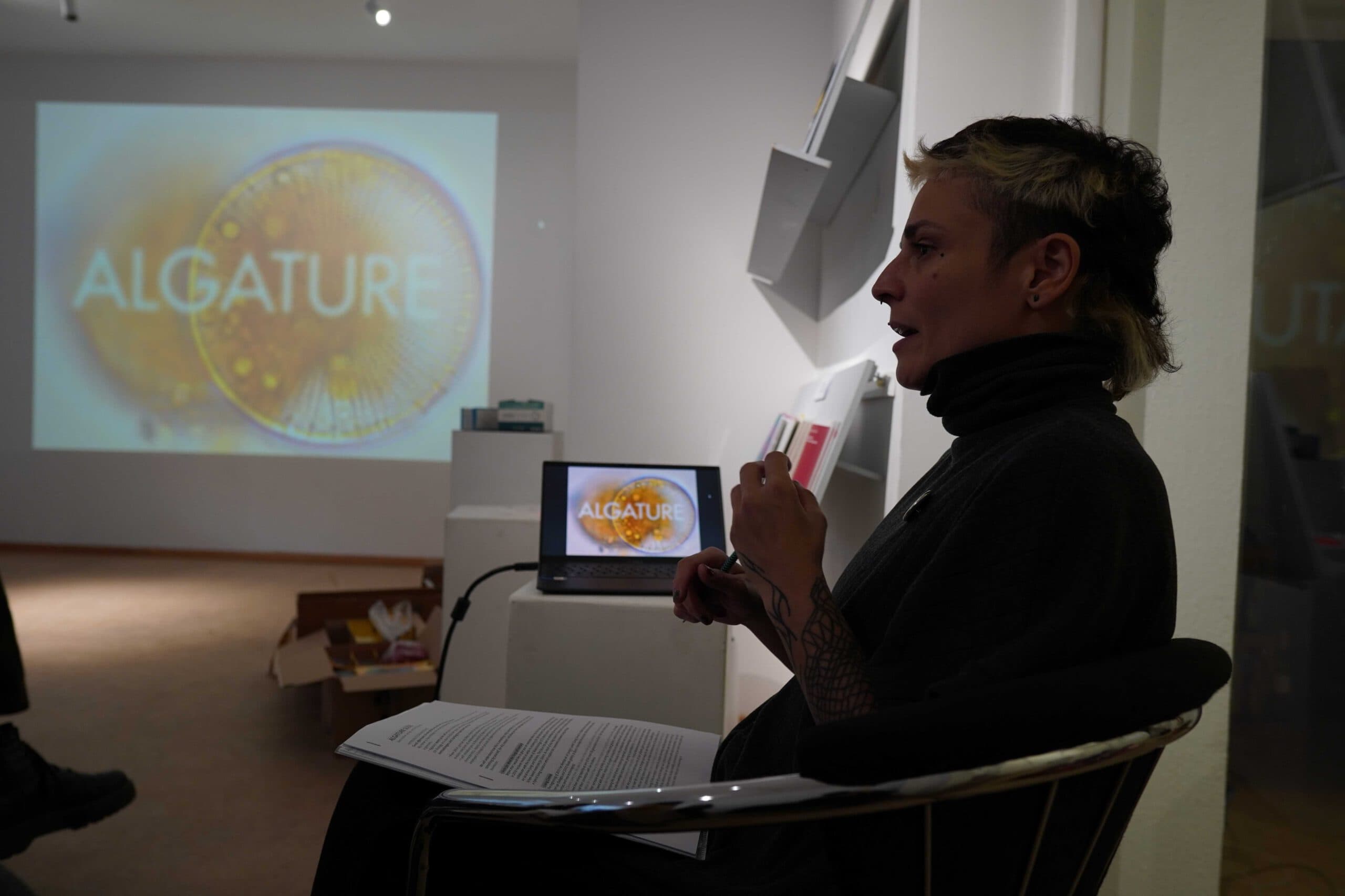 Fara Peluso talking indoors a slide on a wall with the words "Algature" and two diatoms