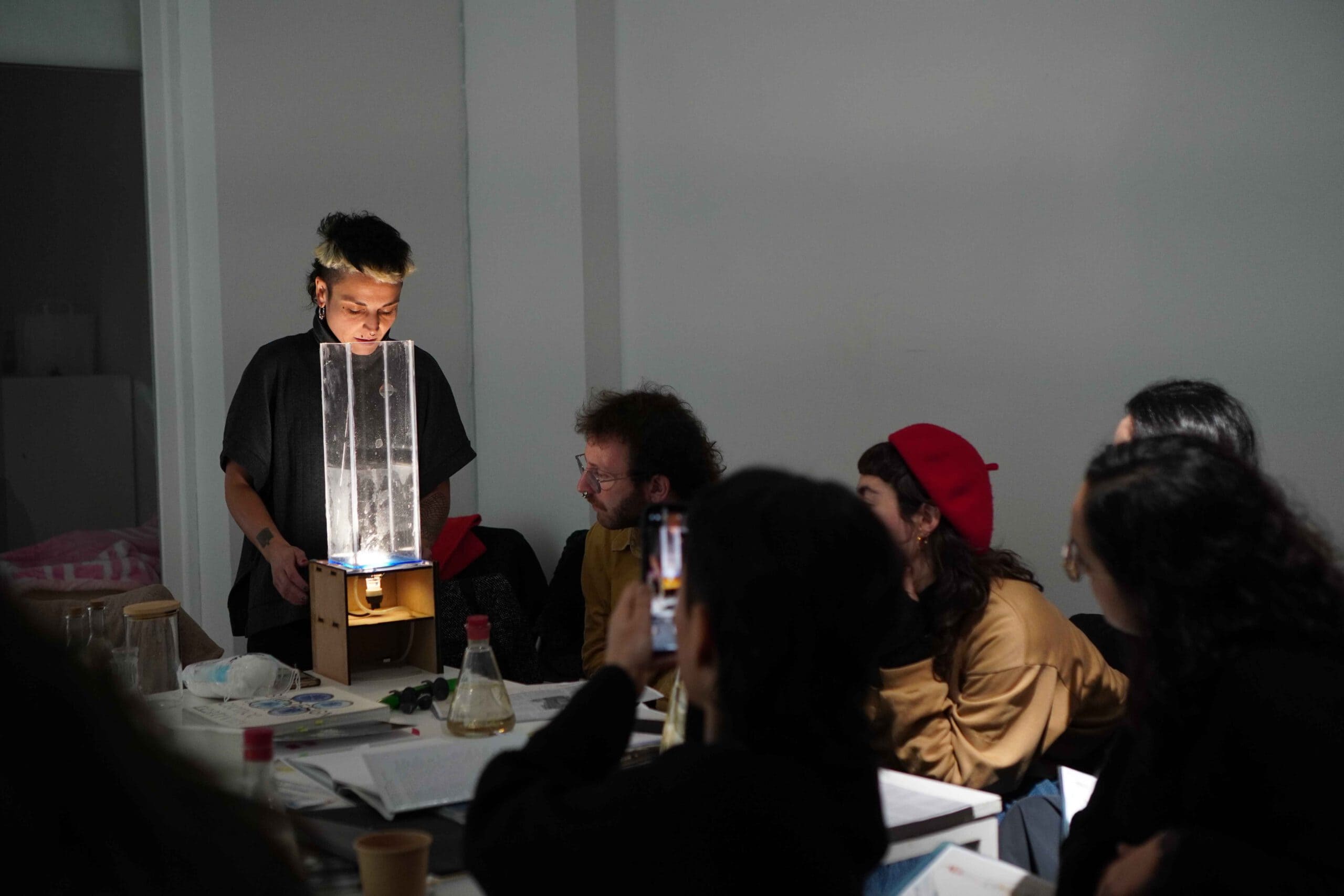 Fara showing a plexiglass container/sculpture to a group of people seated at a table