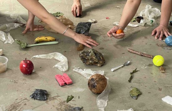 Hands touching and holding objects, a rock, an orange,  on a floor. More objects strewn along the floor.  (This is alt text)