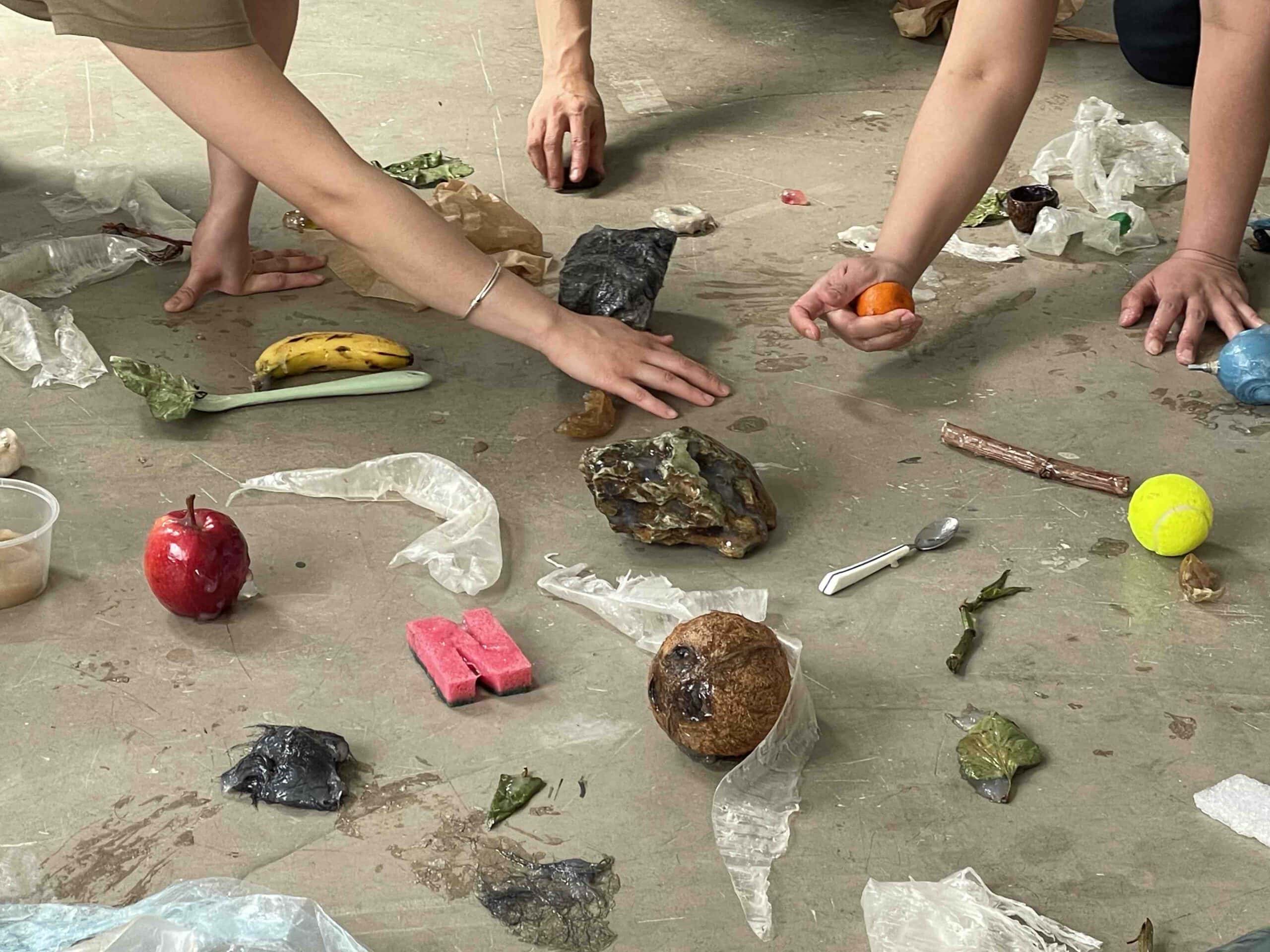 Hands touching objects, rock, an orange, on a floor. covered with natural glue. More objects strewn along the floor.