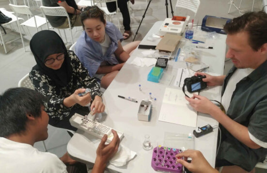 A group of people sitting around a table with laboratory equipment. A woman is pipetting.