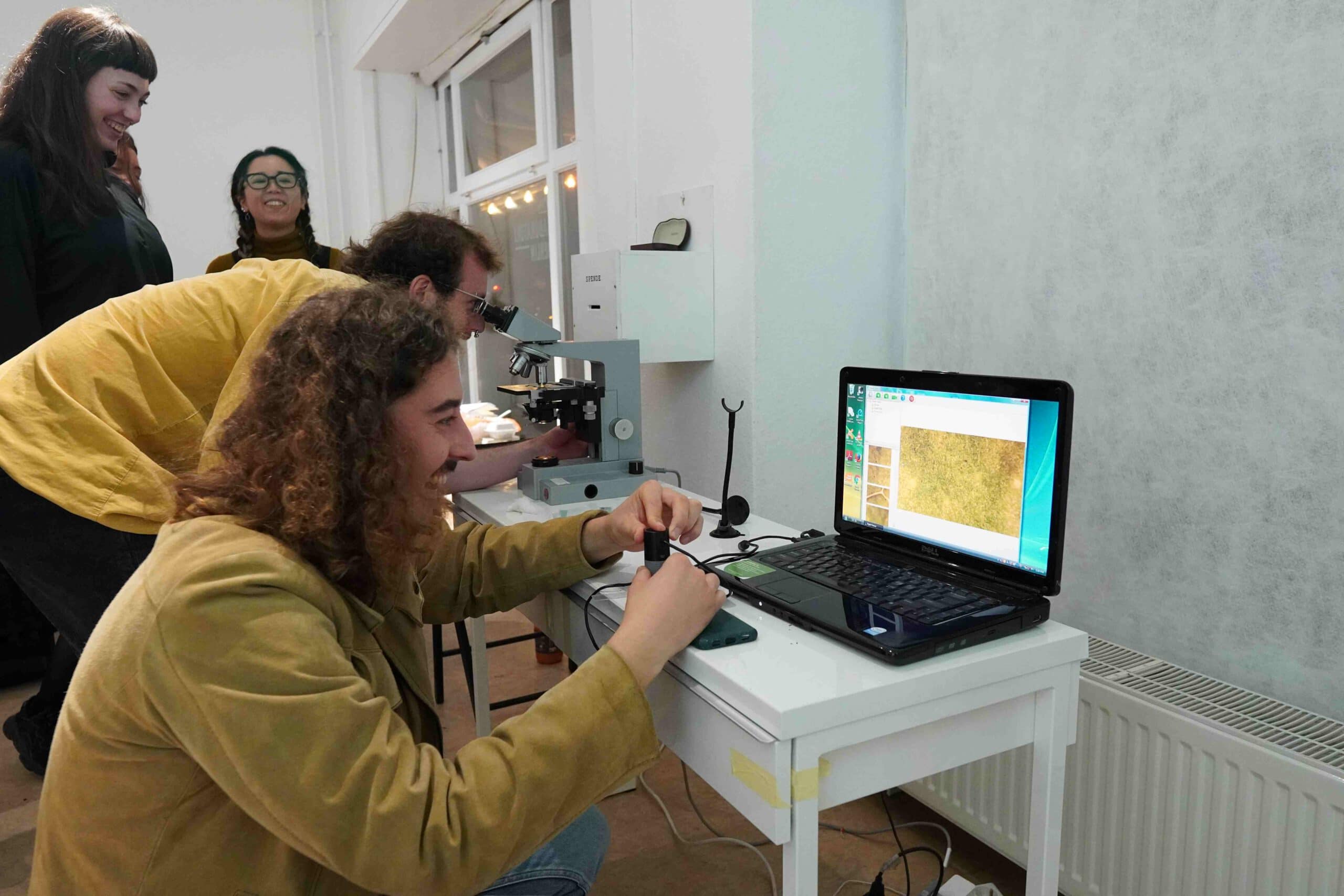 a man smiling and looking at a laptop while using a digital microscope, other people smile or look through microscopes