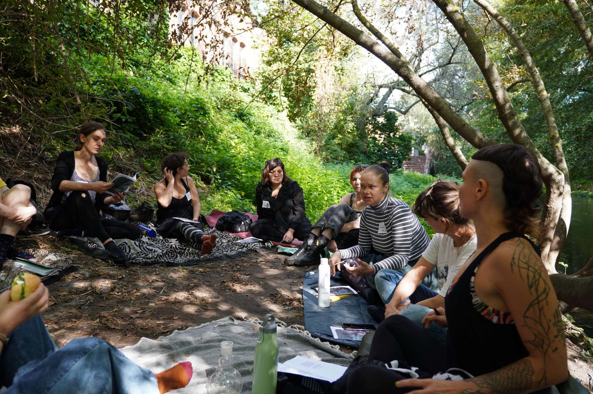 group of people sitting and talking under trees beside a river