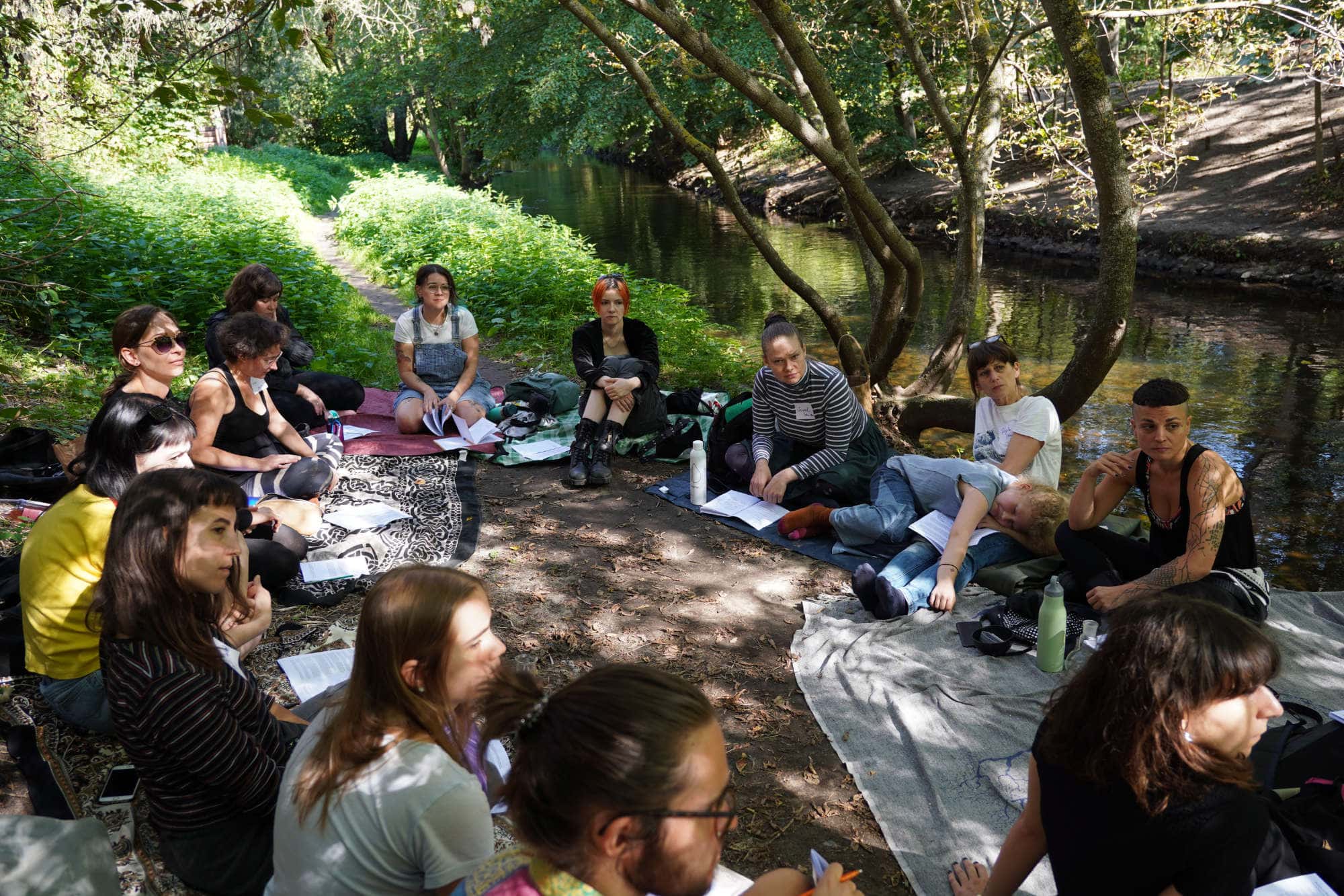 group of people sitting and talking under trees beside a river